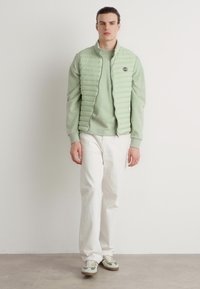 Chaleco acolchado verde claro con acolchado horizontal, superpuesto a un sudadera verde a juego. Combinado con pantalones blancos y zapatillas con detalles verdes.