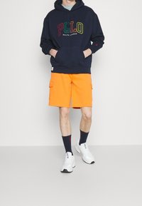 Navy hoodie med flerfärgad "Polo Ralph Lauren"-logotyp, orange shorts med cargofickor, vita sneakers och marinblå strumpor.