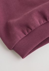 Gros plan d'une manche de sweat-shirt violet montrant le poignet côtelé et la texture douce du tissu tricoté sur une surface claire.