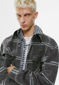 Šedá flanelová košele-jacket s károvaným vzorom, so zapínaním na gombíky vpredu, veľkými vreckami a s kostkovanou košeľou pod ňou.