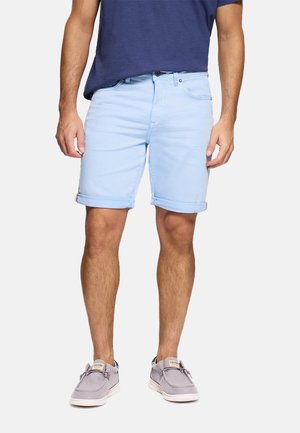 Hombre con pantalones cortos de mezclilla azul claro remangados, camiseta azul marino y zapatos casuales grises de pie sobre fondo blanco.