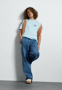 T-shirt azzurro a maniche corte con logo, abbinato a jeans in denim blu larghi e sneakers beige. Maglione appoggiato sulla spalla.
