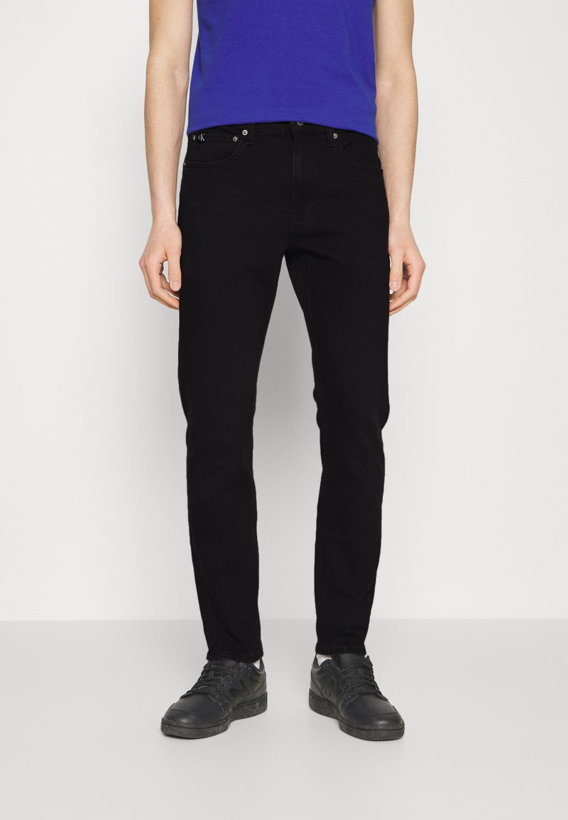 Zwarte denim jeans met een slim fit, een medium taille en een vijf-pocket ontwerp. Gepaard met een blauw T-shirt en zwarte sportschoenen.