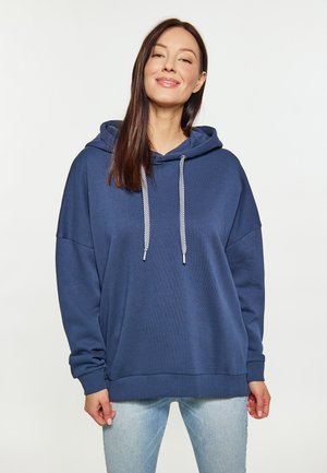 Jonge vrouw die een losse marineblauwe hoodie met witte trekkoorden en lichtblauwe spijkerbroek draagt, staand tegen een effen lichte achtergrond.