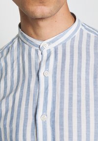 Gros plan sur une personne portant une chemise à rayures verticales bleues et blanches avec un col mao et trois boutons blancs visibles boutonnés.