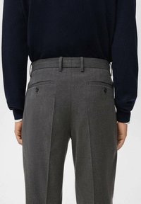 Vedere din spate a unei persoane îmbrăcate cu pantaloni gri din stofă, cu bucle pentru curea și buzunare din spate cu nasturi, asociați cu un top negru cu mânecă lungă.