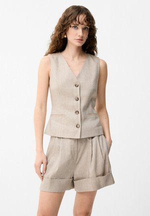 Kvinde iført beige ærmeløs vest med knapper og matchende shorts med opbukket kant, stående med den ene hånd i lommen mod hvid baggrund.