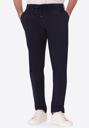 Uomo che indossa pantaloni blu navy con coulisse e sneakers bianche, mani in tasca, in piedi davanti a uno sfondo chiaro e uniforme.