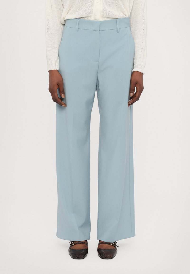 Pantaloni bleu deschis, croiți pe măsură, cu un design cu picioare largi, talie înaltă și pliseuri frontale, asortați cu pantofi maro închis cu accente de cataramă.