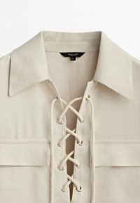 Beige Langarmhemd mit laced Ausschnitt, metallenen Ösen und Schnürung, spitzem Kragen und zwei vorderen Taschen.