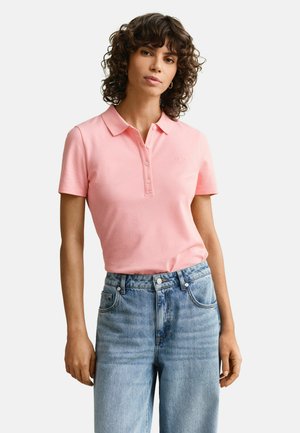 Vrouw met krullend haar die een roze polo met korte mouwen en een hooggetailleerde blauwe jeans draagt, staand tegen een effen achtergrond.