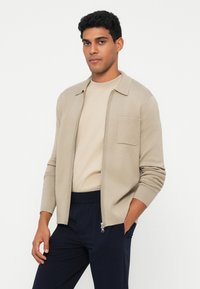 Casaco bege com fecho éclair, colarinho, bolso frontal e punhos canelados. Usado sobre uma camisa bege clara e combinado com calças azul-marinho de cintura elástica.