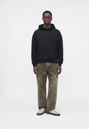 Jeune homme debout, portant un sweat à capuche noir, un pantalon marron ample et des baskets écru, sur un fond clair uni.