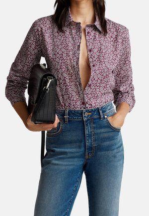 Femme portant une chemise bordeaux à fleurs partiellement boutonnée, jean bleu, tenant un sac à main noir texturé avec une main dans la poche.