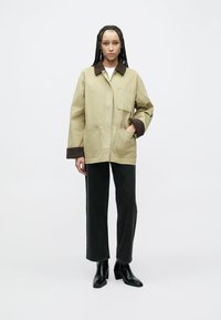 Khaki utiliitjakiga pruun krae, millel on kaks eesmist taskut, kantud valge särgi ja tume teksaga, kokku sobitatud mustade pahkluu-saabastega.