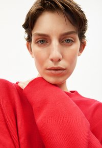 Rode textuurtrui met een losse pasvorm, met ribbels aan de manchetten. Het model is van dichtbij te zien, met een neutrale achtergrond en natuurlijke make-up.