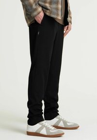 Schwarze Hose aus weichem Stoff mit schmalem Schnitt, ausgestattet mit Seitentaschen. Kombiniert mit weißen Sneakers, die graue Akzente und braune Sohlen haben.