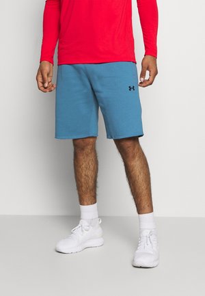 Homme portant un short bleu Under Armour, des chaussures de sport blanches, des chaussettes blanches et une chemise rouge à manches longues, debout devant un fond uni.