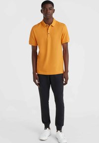 Camisa polo naranja hecha de algodón con cuello tipo solapa, mangas cortas y detalle del logo. Combinada con pantalones negros ajustados y zapatillas blancas.