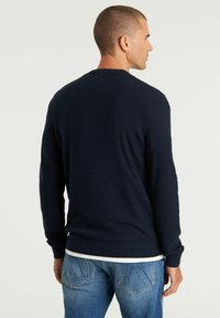 Dunkelblauer Strickpullover mit langen Ärmeln, Rundhalsausschnitt, gerippten Manschetten und Saum. Über einem weißen Shirt getragen. Verfügt über eine subtile Textur.