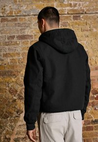 Giacca nera con cappuccio, dal tessuto morbido e polsini a coste, con un colletto foderato in pile, mostrata su uno sfondo di muro di mattoni.