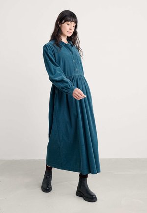 Teal Baumwollcord-Kleid mit langen Ärmeln, Knopfverschluss und Taillennaht, ausgestattet mit einem fließenden Rock und enganliegenden Manschetten, kombiniert mit schwarzen Knöchelstiefeln.