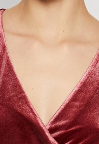 Gros plan sur le haut de la poitrine d'une personne portant un top en velours à décolleté en V profond de couleur rose foncé. La peau présente de petites imperfections naturelles.