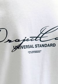 Bílá tkanina s černým potiskem textu "Projekt E... UNIVERZÁLNÍ STANDARD" a menším textem "AGENTURNÍ ZASTOUPENÍ VŠECHNA PRÁVA VYHRAZENA."