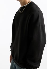 Personne portant un sweat-shirt noir surdimensionné, un collier de perles et un jean bleu, debout contre un fond blanc.