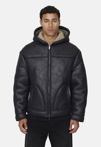 Giacca nera imbottita con finitura texturizzata, dotata di cappuccio e chiusura con zip. Foderata in pile beige chiaro per il calore. I dettagli delle cuciture sono visibili.