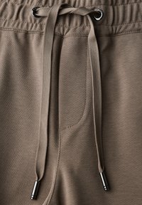 Beige stoffen sweatpants met een elastische tailleband, twee metalen oogjes en een bijpassende trekkoord met metalen uiteinden.