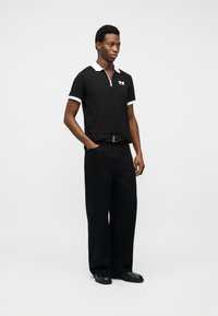 Νέος άνδρας που στέκεται με το ένα χέρι στην τσέπη, φορώντας μαύρο πόλο με λευκό γιακά και λεπτομέρειες, μαύρο παντελόνι φαρδύ και μαύρα παπούτσια.