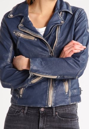 Personne portant une veste en cuir bleue zippée avec des fermetures éclair et des boutons en argent, les bras croisés sur la poitrine, associée à un jean foncé.