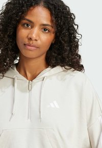 Light gray zip-up hoodie made of soft fabric, featuring a front pocket and white logo on the left chest. Textured, relaxed fit design.