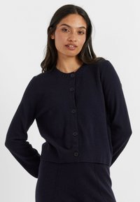 Mariniereblauwe cardigan met een ronde halslijn, ribbels aan de manchetten en onderkant, en zes grote zwarte knopen aan de voorkant. Zacht, gestructureerd breimateriaal.