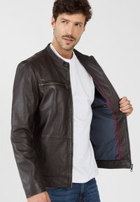 Dunkelbraune Lederjacke mit Stehkragen, Frontreißverschluss und Brusttasche. Innenfutter in Blau mit roten Akzenten. Lässige Passform.