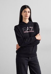 Fekete kapucnis pulóver, rajta reflektáló felirattal: "EA7 EMPORIO ARMANI". A modell karjait összefonva pózol egy semleges háttér előtt.