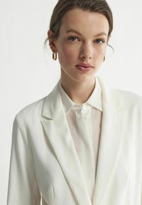 Biała lniana marynarka z klapami, w zestawieniu z lekką, przezroczystą koszulą na guziki. Złote kolczyki w kształcie kół podkreślają całość.