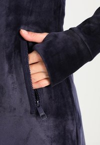 Una mano inserita parzialmente nella tasca con zip di una giacca in peluche blu scuro con maniche lunghe.