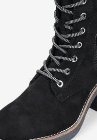Stivale alla caviglia in suede nero caratterizzato da un design alto con lacci, cuciture bianche a contrasto e una suola in gomma testurizzata. I lacci sono neri e bianchi.