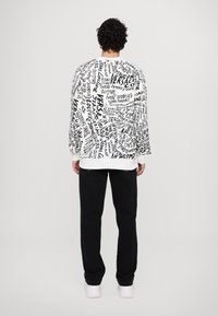 White sweatshirt with black graphic text detail, featuring a relaxed fit and ribbed cuffs, paired with black pants and white shoes.