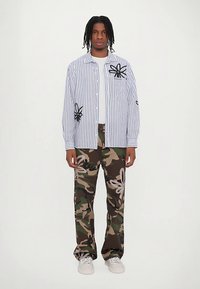 Moški stoji v belem črtastem srajcu z črnimi cvetličnimi vzorci, vojaških hlačah s cvetličnimi motivi in belih športnih copatih na bežni podlagi.