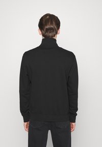 Musta huppari, jossa on korkea kaulus, pitkät hihat ja ribbaatut hihansuut. Sileä pinta, yksinkertainen muotoilu ja vartalonmyötäinen vyötäröllä.