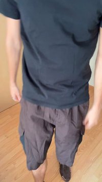 Crna majica u kombinaciji s tamnim cargo šorcem s bočnim džepovima. Materijal izgleda lagano. Pod je drven, stvarajući neutralnu pozadinu.