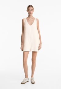 Helles beige ärmelloses Kleid mit V-Ausschnitt und lockerer Passform, aus strukturiertem Stoff; kombiniert mit lässigen beige-weißen Sneakern.