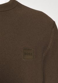 Brun texturerad tröja med ribbad rund halsringning och en fyrkantig lapp med "BOSS"-logotypen på vänstra bröstet.