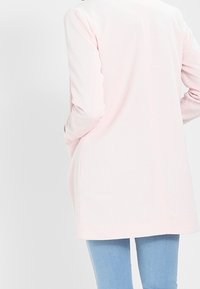 Light pink long-sleeve cardigan with a straight cut, textured fabric, open front, and side pockets, paired with light blue jeans.