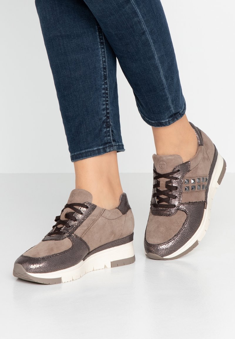 Bruna mocka och metalliska sneakers med texturerade detaljer, snörningsdesign och en något upphöjd vit sula. Har nitar på sidan.