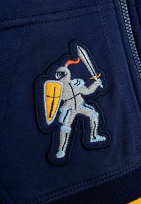 Un tejido azul marino presenta un detallado diseño bordado de un caballero, sosteniendo una espada y un escudo, con acentos en amarillo y naranja.