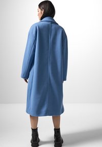 Lungo cappotto di lana blu con collo a pistagna, spalle basse e cuciture laterali. Rifinito con un'orlo dritto e indossato con stivaletti neri fino alla caviglia.